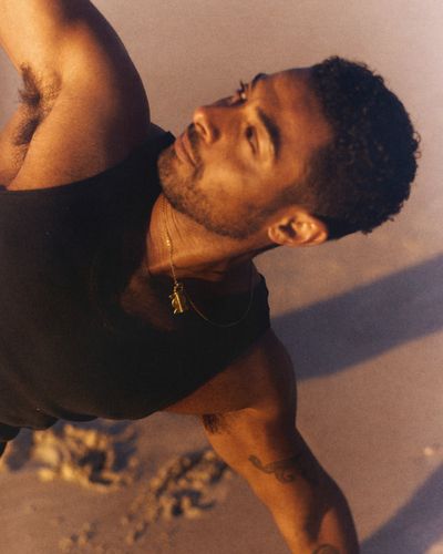 A man in a dark singlet holds a side stretch on the sand in late afternoon light, looking upward.