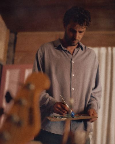 A man in a grey shirt mixes paint on a wooden palette in a timber room, with a guitar out of focus in the foreground.